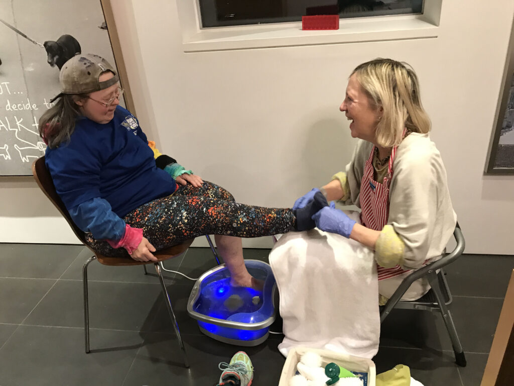 Franke James giving Teresa Heartchild a foot spa. Both sisters are laughing. Teresa is wearing floral leggings and a blue shirt. Franke is wearing a beige sweater and a red striped apron.