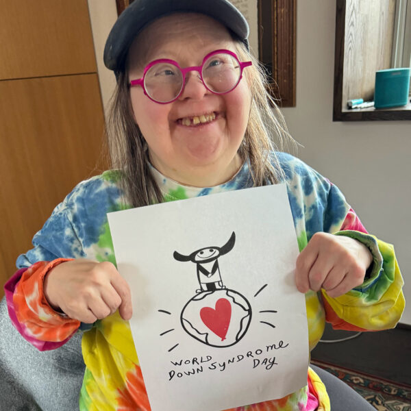 Teresa Heartchild, an artist with Down syndrome, is wearing pink glasses, a backwards baseball cap and smiling. She is holding up her design for the T-shirt. It's a small black and white figure with horns on her head standing on top of the world. The globe has a large red heart in it. Below it, Teresa has written World Down Syndrome Day. Photo by Franke James.