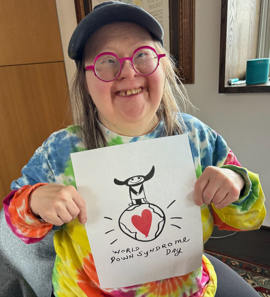 Teresa Heartchild, an artist with Down syndrome, is wearing pink glasses, a backwards baseball cap and smiling. She is holding up her design for the T-shirt. It's a small black and white figure with horns on her head standing on top of the world. The globe has a large red heart in it. Below it, Teresa has written World Down Syndrome Day. Photo by Franke James.