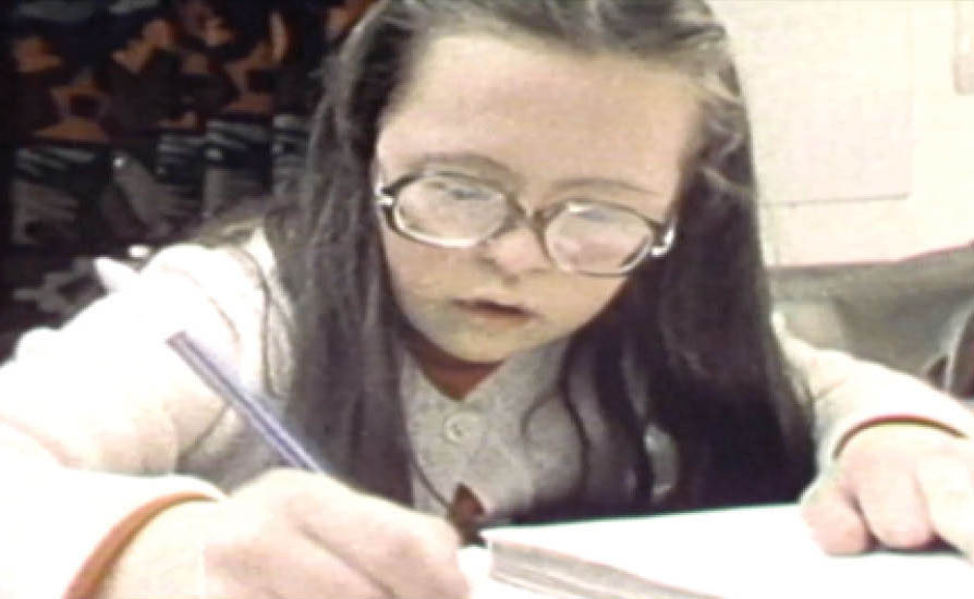 Teresa Heartchild at age 13 in a still from the documentary Exploding the Myth aired in 1979 shows Teresa writing in her notebook. She is white and has long brown hair and glasses. She has Down syndrome. Images and text used with permission from Community Living Ontario
