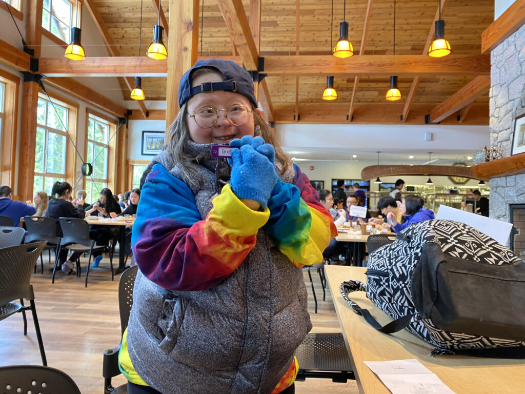Teresa Heartchild at the 2020 Inclusion BC retreat for people with developmental disabilities and their families to learn about rights. Teresa is smiling at the camera and holding her badge up. She is a white woman with Down syndrome wearing round wire glasses, a grey down vest, a tie-dye sweatshirt, and a backwards ball cap. Photo by Franke James