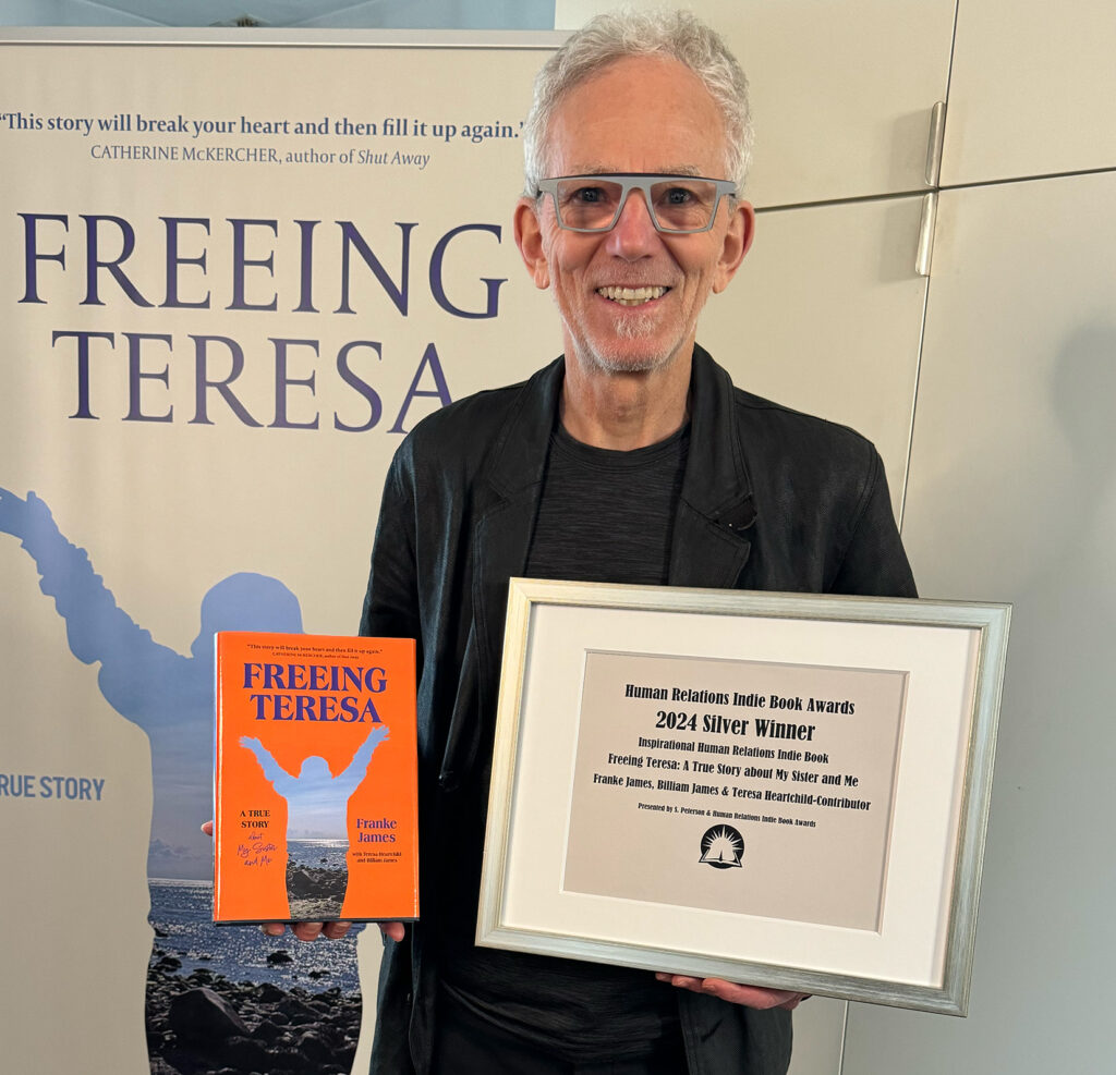 Billiam James is smiling and holding the framed 2024 Silver award for Inspirational from the Human Relations Indie Book Awards. Billiam is wearing a black sports jacket and black t-shirt. Photo by Franke James.