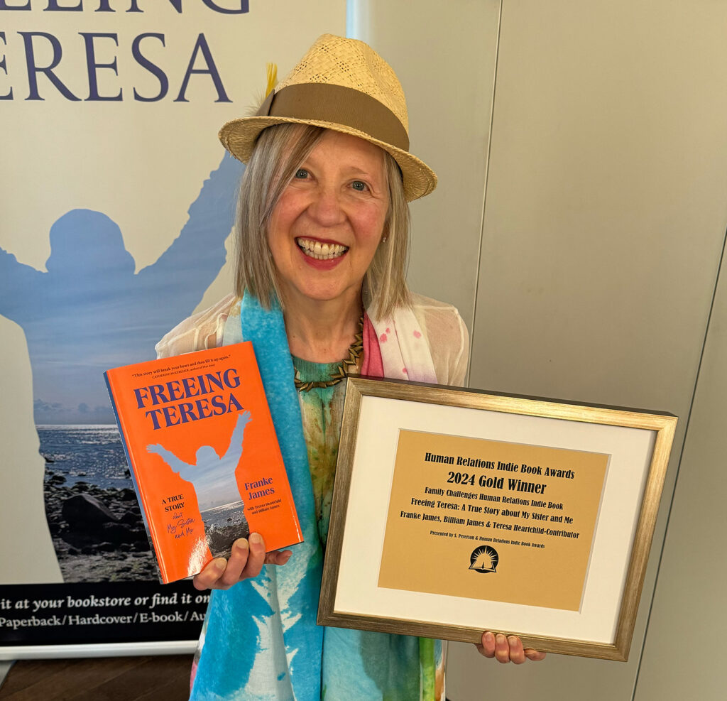 Franke is smiling and holding up the framed 2024 Gold award for Family Challenges from the Human Relations Indie Book Awards. Franke is wearing a painterly pastel dress, matching scarf and straw hat. Photo by Billiam James.