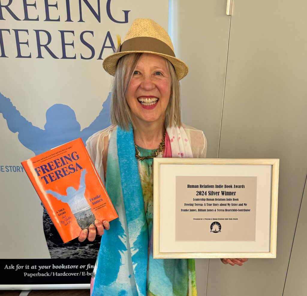 Franke is smiling and holding the framed 2024 Silver award for Leadership from the Human Relations Indie Book Awards. Franke is wearing a painterly pastel dress, matching scarf and straw hat. Photo by Billiam James.
