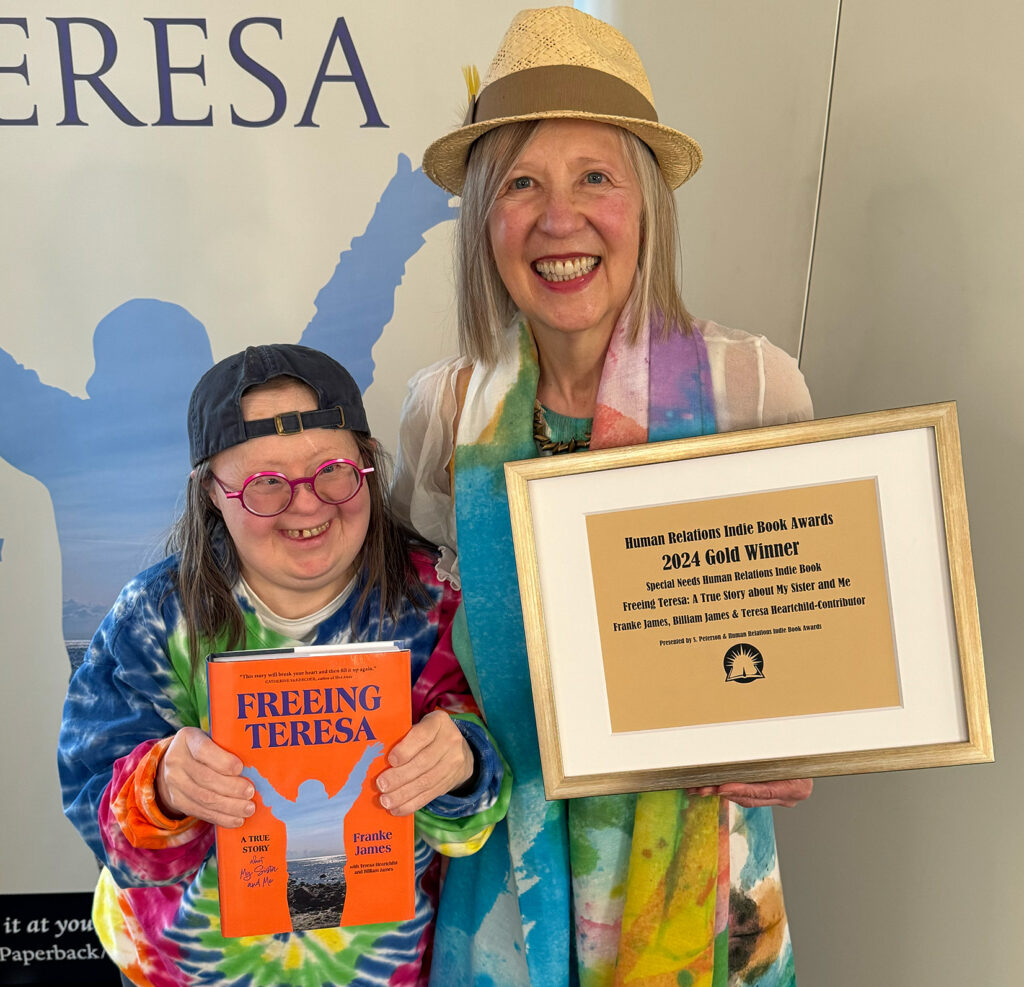 Franke James, the author of Freeing Teresa stands beside her sister Teresa Heartchild who has Down syndrome and is a contributor to Franke's book. Teresa is wearing pink glasses, a tie-dye shirt, a backwards ball cap and smiling. Franke is smiling and holding up the framed 2024 Gold award for Special Needs from the Human Relations Indie Book Awards. Franke is wearing a painterly pastel dress, matching scarf and straw hat. Photo by Billiam James.