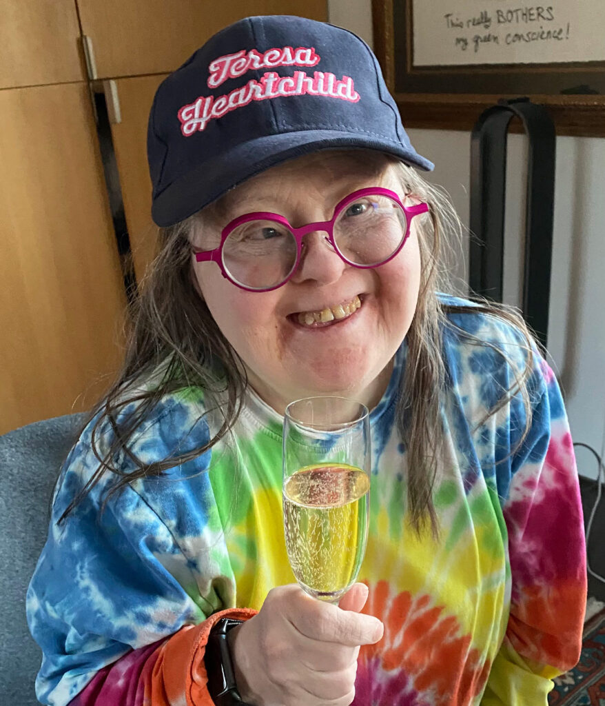 Teresa celebrated the ten year anniversary with a glass of champagne! She is wearing pink glasses, a tie-dye shirt and ball cap