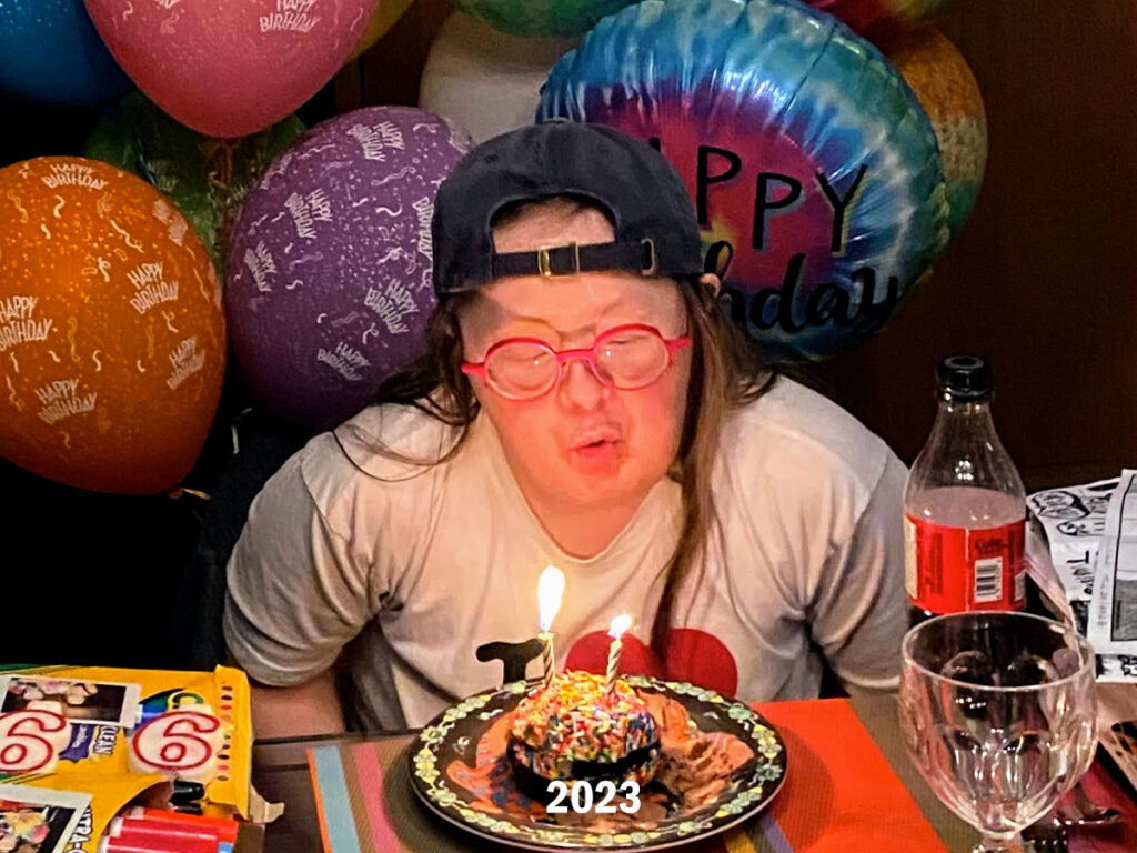 Teresa leans forward and blows out the candles on her 2023 birthday cake. She is surrounded by colourful helium birthday balloons.